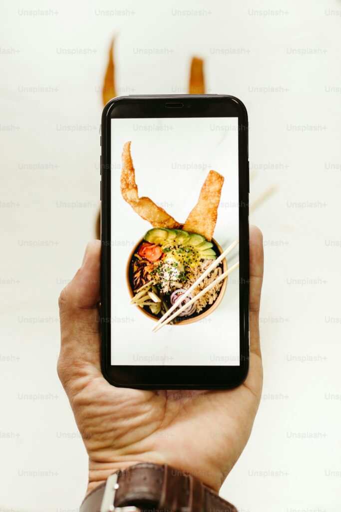 A hand holding a smartphone displaying an image of a bowl of ramen with various toppings and two pieces of fried dough on a light background.