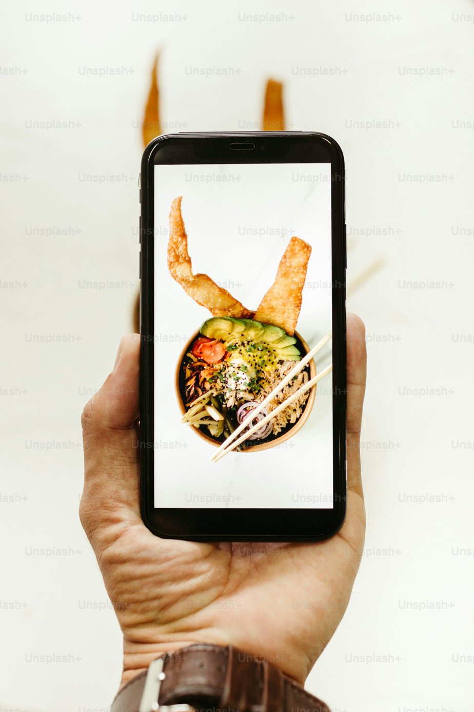A hand holding a smartphone displaying an image of a bowl of ramen with various toppings and two pieces of fried dough on a light background.