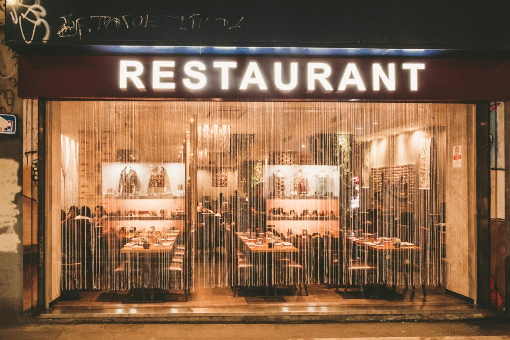 Exterior view of a warmly lit restaurant with curtains, showing neatly arranged tables and chairs inside.