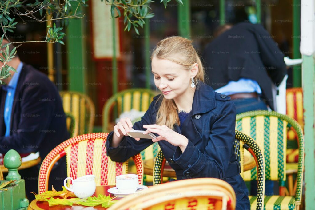 A woman in a dark coat sits at an outdoor café table, taking a photo of food with her phone. There are cups of coffee on the table and colorful chairs around her.